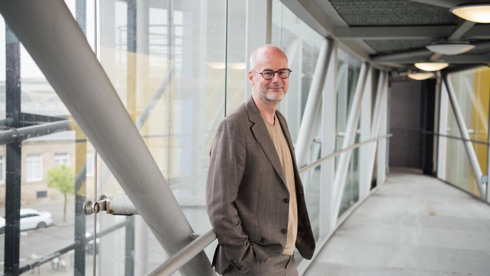 Professor Volker Straub standing in a windowed hallway, smiling at the camera