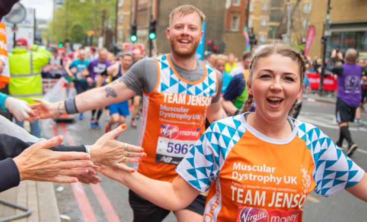 A young female runner accompanied by a young male runner, high five spectators while running the Virgin London Marathon in their MDUK t-shirts.