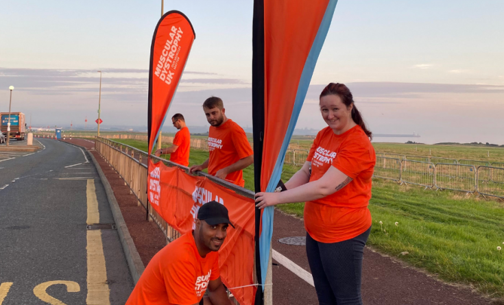 Jodie is tying an MDUK flag to the railing and is smiling at the camera. Krish is opposite Jodie, crouched on the floor tying it to the bottom of the railing.
