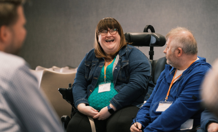 A woman in a powerchair sits laughing, she converses with a man to her right.
