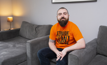 Joe is sat on a footstall in his living room, wearing an orange T-shirt that reads 'Straight outta dystrophin' in all-caps. 