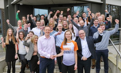 Staff from Bidwells head office are all stood in front of the building with one arm in the, cheering. At the front is Matt Allen and Amy O'Hanlon shaking hands