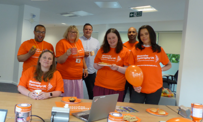 Image of 7 volunteers in their MDUK Orange T-shirts holding cakes, balloons and in front of a table of fundraising materials