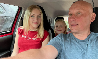 The photo is taken inside a red car. Dean sits on the passenger side with his daughter Grace, an 18 year old blonde girl in the drivers seat. A Younger blonde girl, Millie, sits behind the driver.