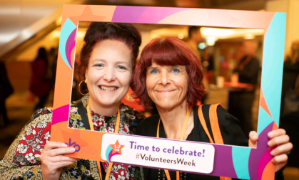 Two women pose with a cardboard photo frame that reads "Time to celebrate! #VolunteersWeek"