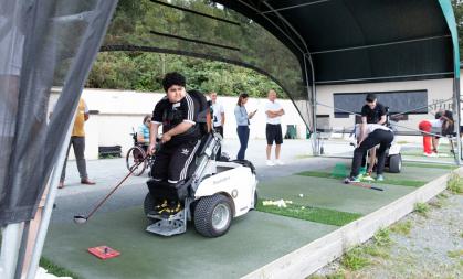 A young boy is stood up, using assistive technology, about to hit a golf ball in a driving range. In the background, someone is doing the same.