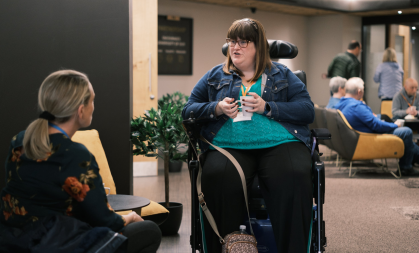 A woman in a wheelchair sits with a mug of tea. She talks to another woman whose back is to the camera.