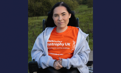 Lauren sits in her powerchair in a field. She faces the camera head on. She wears an orange Muscular Dystrophy UK t-shirt.