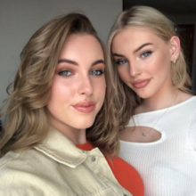 A selfie with two women. The one on the left of frame is brown haired, blue eyed with an orange top on. The one on the right is blonde haired, blue eyed with a white top on. Both look into the camera.