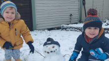 Two boys are crouching in the snow next to their snowman. They are wearing puffer coats, woolly hats, gloves and wellies. The snowman is hearing a woolly hat too.