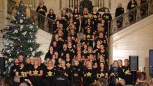 A choir stand on the stairs of a great hall. They sing to an audience next to a large Christmas tree. 