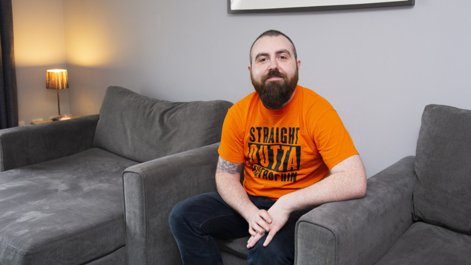 Joe is sat on a footstall in his living room, wearing an orange T-shirt that reads 'Straight outta dystrophin' in all-caps. 