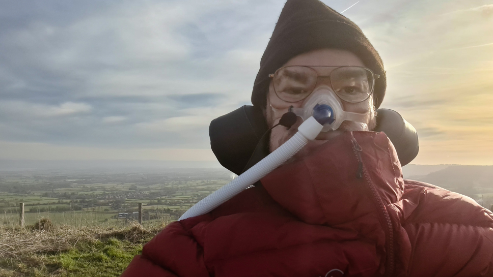 Image of Daniel in front of a scenic background of countryside and a cloudy, blue sky. Daniel is wearing a red puffer jacket, glasses, hat and his CPAP mask.