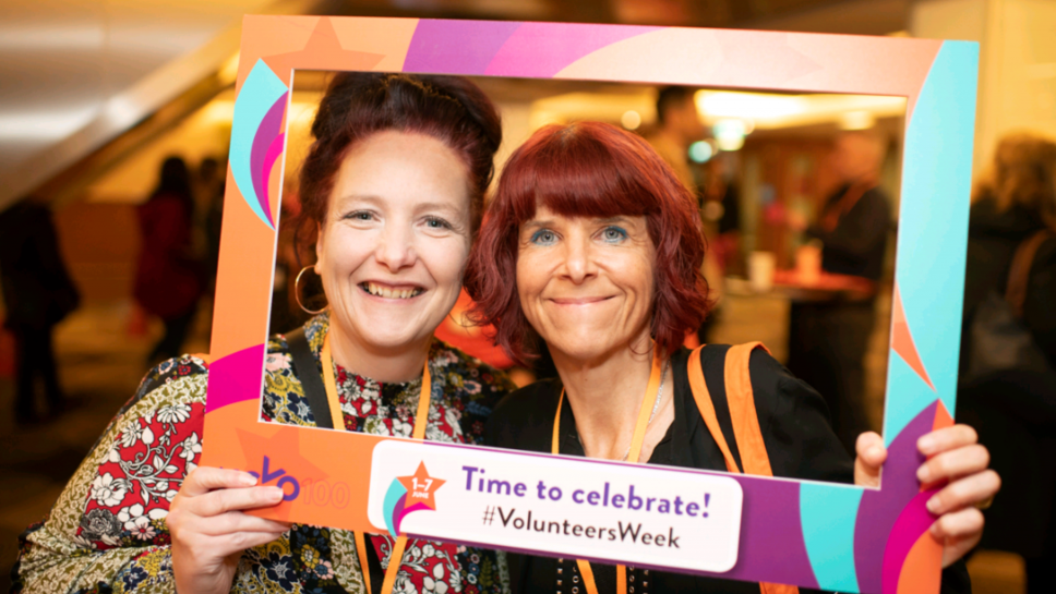 Two women pose with a cardboard photo frame that reads "Time to celebrate! #VolunteersWeek"